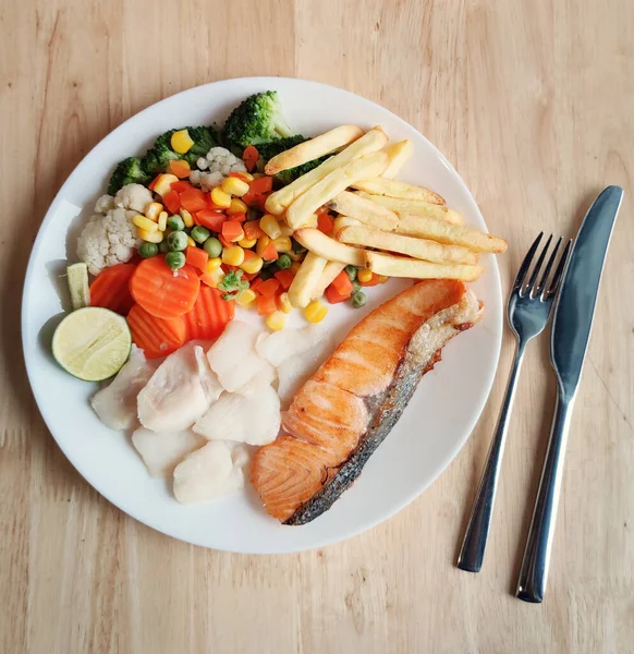 Grilled salmon fish and baked cod fish decorated with various vegetables corn carrot peas lemon French fries and sauces on white flat plate table on wooden table, food stylist for international fresh market restaurant, menu advertising backgrounds — Stock Image Grilled Salmon Fish Baked Cod Fish Decorated Various Vegetables Corn Stock Photo