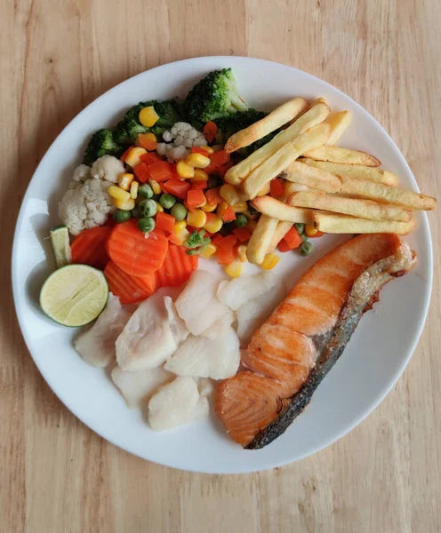 Grilled salmon fish and baked cod fish decorated with various vegetables corn carrot peas lemon French fries and sauces on white flat plate table on wooden table, food stylist for international fresh market restaurant, menu advertising backgrounds — Stock Image Grilled Salmon Fish Baked Cod Fish Decorated Various Vegetables Corn Stock Photo