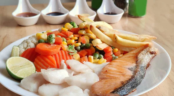 Grilled salmon fish and baked cod fish decorated with various vegetables corn carrot peas lemon French fries and sauces on white flat plate table on wooden table, food stylist for international fresh market restaurant, menu advertising backgrounds — Stock Image Grilled Salmon Fish Baked Cod Fish Decorated Various Vegetables Corn Royalty Free Stock Images