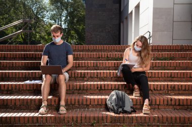 Coronavirus salgını sırasında üniversiteye dönmek. Maskeli iki üniversite öğrencisi merdivenlerde oturmuş sosyal mesafeyi koruyorlar..