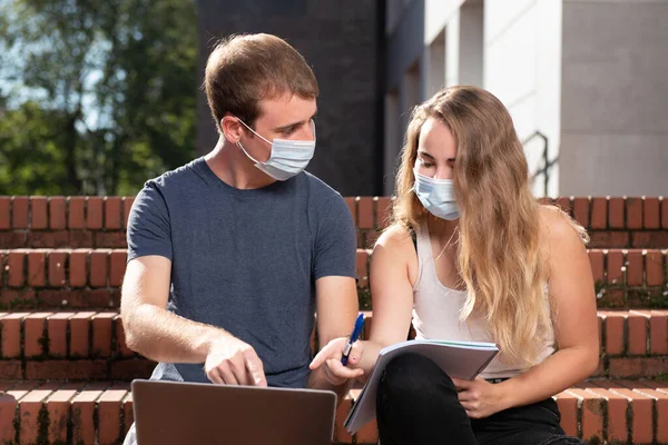 Coronavirus salgını sırasında üniversiteye dönmek. Maskeli iki üniversite öğrencisi merdivenlerde otururken dizüstü bilgisayarı işaret ediyor..