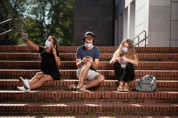 Sosyal uzaklık kavramı. Merdivenlerde oturan üç üniversite öğrencisi ve yüz maskesi takarak akıllı telefonlarını kullanıyorlar ve koronavirüs salgını sırasında sosyal mesafeyi koruyorlar..