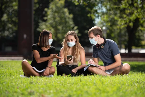 Coronavirus ya da Covid-19 sırasındaki birlikteliğe. Üç üniversite öğrencisi parkta oturmuş maske takmış sohbet ediyorlar..