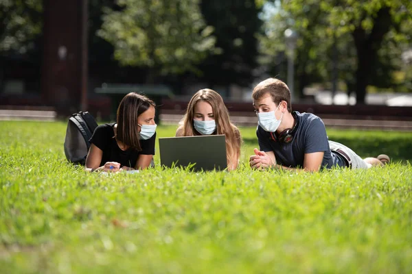 Coronavirus ya da Covid-19 sırasındaki birlikteliğe. Üç üniversite öğrencisi, dizüstü bilgisayarlarıyla parkta yüz maskesi takıp sohbet ediyorlar..