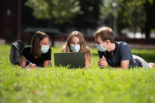 Coronavirus ya da Covid-19 sırasındaki birlikteliğe. Üç gülümseyen üniversite öğrencisi, dizüstü bilgisayarlarıyla parkta yüz maskesi takıp sohbet ediyorlar..