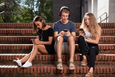 Dijital teknoloji bağımlılığı. Merdivenlerde oturan ve akıllı telefonlarına odaklanmış üç genç öğrenci..