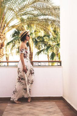 Moda gözlüklü, şapkalı ve kumsalda uzun elbiseli genç bir kadın. 
