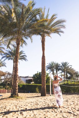 Uzun elbiseli ve şapkalı bir kadın okyanusun kenarında kumda yürüyor. 