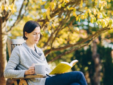 Sıcak örgü giyen kadın elbise bir fincan sıcak çay ya da kahve içme ve güneş ışığı - sezon, edebiyat, boş zaman zaman ve insanların kavramı - açık havada bir kitapta okuma güzel sabah bahçedeki