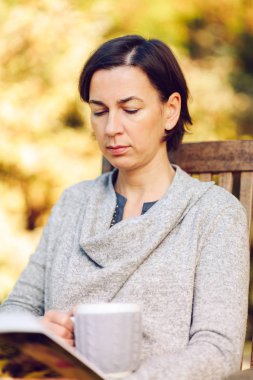 Sıcak örgü giyen kadın elbise bir fincan sıcak çay ya da kahve içme ve güneş ışığı - sezon, edebiyat, boş zaman zaman ve insanların kavramı - açık havada bir kitapta okuma güzel sabah bahçedeki