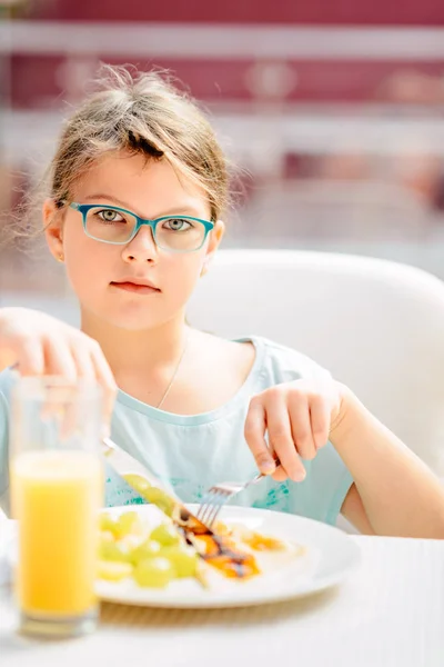 Menina alegre comer panquecas, frutas frescas e beber laranja — Fotografia de Stock