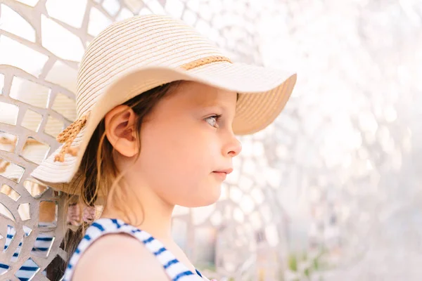 Маленькая задумчивая девочка в соломенной шляпе, стоящая на фоне яркой мерцающей стены — стоковое фото