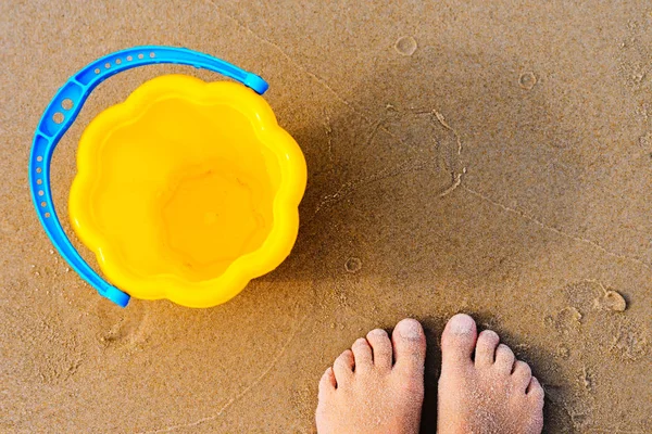 Die nackten Füße des Babys an einem goldenen Sandstrand neben einem gelben Spielzeugeimer — Stockfoto