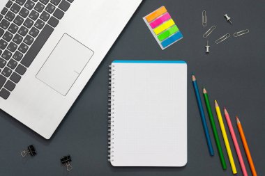 Ofisteki işyerinde. Gri masa. Fotoğrafta bir dizüstü bilgisayar, renkli çıkartmalar, kalemler, ofis aksesuarları var. Defterin yanında çok renkli kalemler var. Ofis geçmişi.