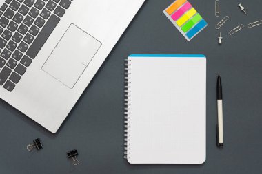 Ofisteki işyerinde. Gri masa. Fotoğrafta bir dizüstü bilgisayar, renkli çıkartmalar, kalemler, ofis aksesuarları var. Defterin yanında çok renkli kalemler var. Ofis geçmişi.