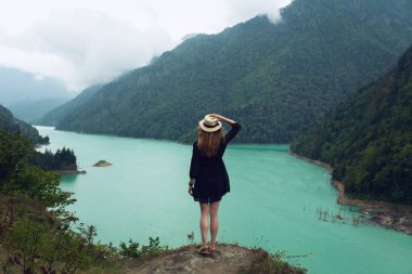 Şapkalı ve elbiseli bir kız Svaneti 'deki Ingur Barajı' nın arka planında bir taşın üzerinde duruyor. Yazın Kafkas Dağları