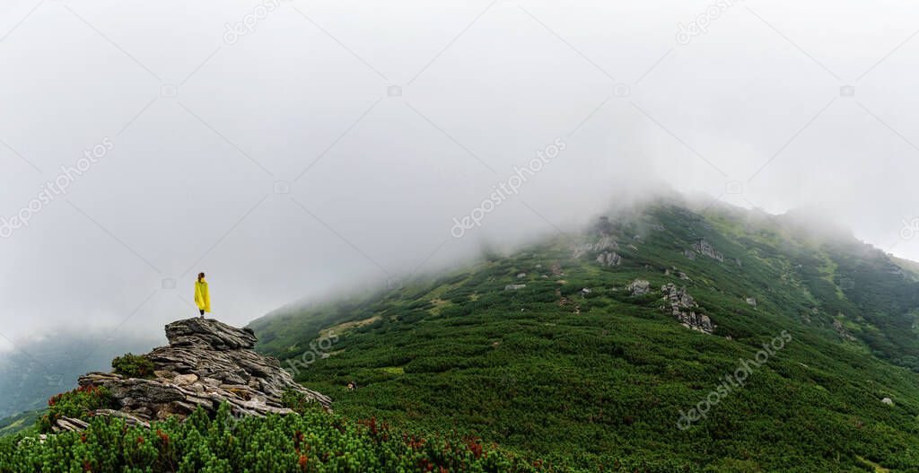 A girl in a yellow raincoat stands on top of a stone. Foggy mountain range in the Ukrainian Carpathians. Rainy day in the mountains.