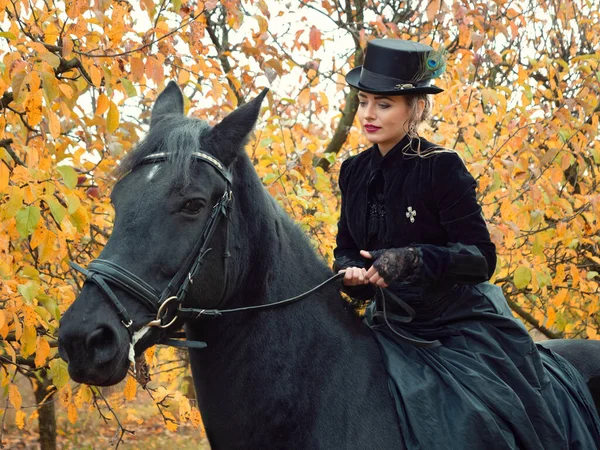 Menina em um vestido preto montando um cavalo preto — Fotografia de Stock