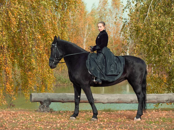 Mädchen im schwarzen Kleid reitet auf einem schwarzen Pferd — Stockfoto