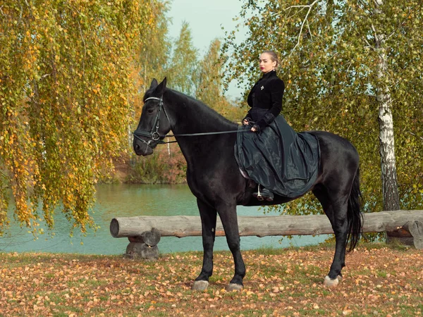 Девушка в черном платье верхом на черной лошади — стоковое фото