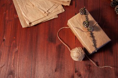 Tahta bir yüzeyde, köknar kozalaklarıyla süslenmiş el yapımı bir karton kutu. Yakınlarda hediye paketinin yapıldığı kağıt poşetler var. Sıfır atık. Boşluğu kopyala