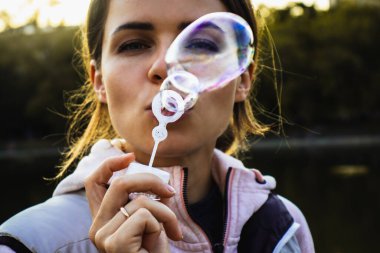 Genç ve güzel kadın dışarıda köpükler saçıyor. O, bir çocuk olarak zamanın tadını çıkarıyor. Fotoğraf modeli sabun köpüklerinin arasından bakıyor..