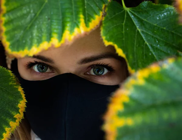 The portrait of the woman with leaves and mask. A young woman uses the black mask like a ninja-style photo session partly covers her face by green tree leaves