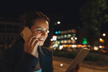 Modern woman multitasking with smartphone and digital tablet on city street at night