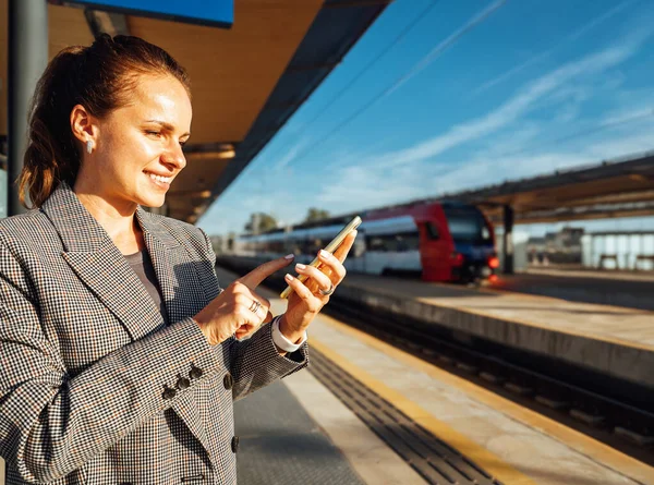 Smiling woman commuter checking mobile phone on railroad station platform.