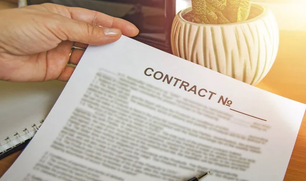 Close-up of a womans hand holding a paper business contract, office background, glow on the background of a laptop, flower, table — Stock Photo, Image