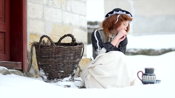 Red-haired girl sits near a village house in winter. Cinderella is tired of hard housework — Stock Video