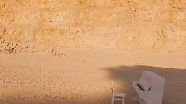 Piano blanc et une chaise blanche se tiennent sur un sable jaune sur le fond des rochers. Intéressante histoire. Vue aérienne — Video