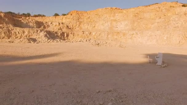 Young man in ethnic clothes barefoot approaches the white piano against the backdrop of a sand quarry. Aerial view — Stock Video