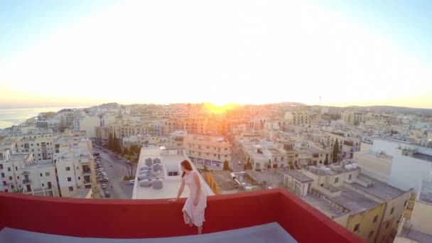 美しい少女は、都市の背景の屋根の上に座っています。写真家のためにポーズの女の子。航空写真ビュー — ストック動画
