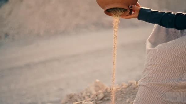 Chica en vestido étnico se sienta cerca de la carretera y vierte arena de la jarra. El agua se convirtió en arena — Vídeos de Stock