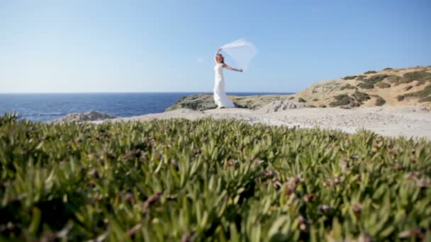 Een gelukkige bruid staat op de kust onder een bruidssluier wapperen in de wind. Het concept van een gelukkig huwelijk en een gelukkig gezinsleven — Stockvideo