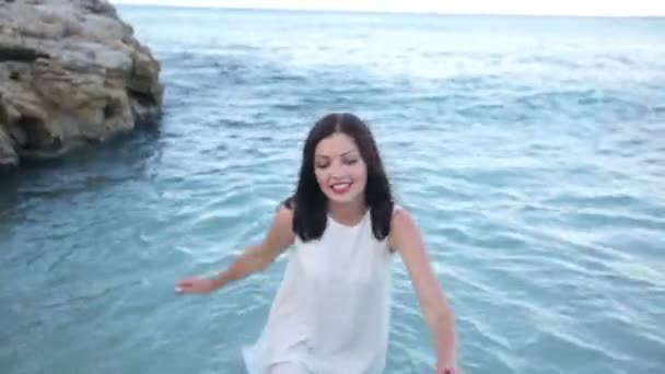 Hermosa mujer morena joven sexy en un vestido mojado blanco en la orilla del mar en un claro día soleado de verano — Vídeo de stock