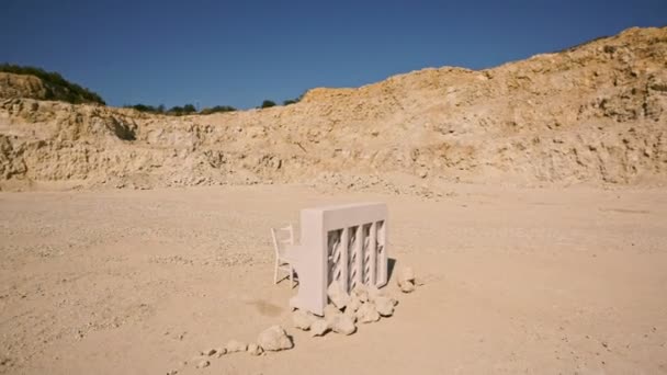 Piano branco e uma cadeira branca ficam em uma areia amarela no fundo das rochas ao pôr do sol. Enredo surreal. Vista aérea — Vídeo de Stock