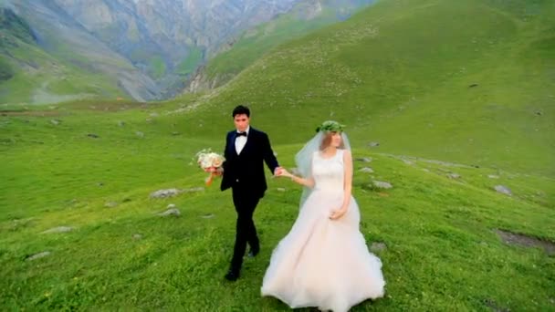 Verliefd jonggehuwden lopen in de avond in de weide tegen de achtergrond van prachtige bergen — Stockvideo
