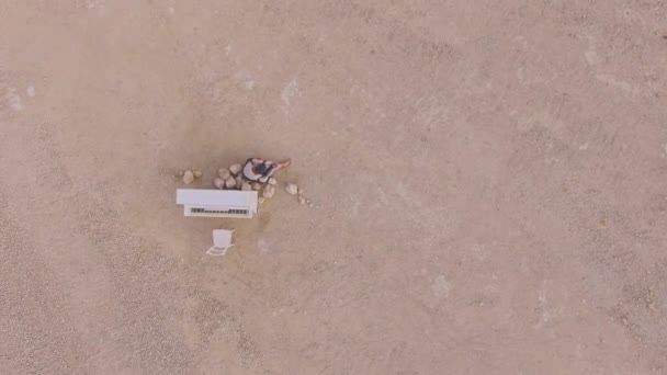 Jonge barefoot vrouw zitten in de woestijn in de buurt van een witte piano. Bekijk van bovenaf. Surrealistisch verhaallijn. Luchtfoto. — Stockvideo