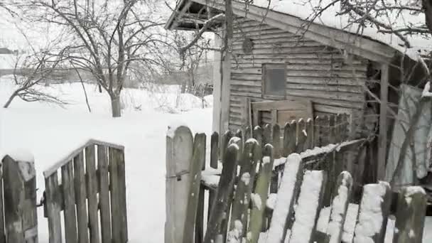 Ένα παλαιό ερειπωμένο ξύλινο σπίτι με ένα φράχτη ξεχαρβαλωμένος και έναν οπωρώνα κοντά το χειμώνα. — Αρχείο Βίντεο