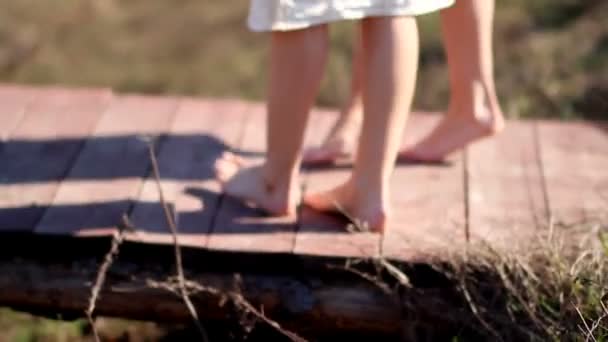 Дві дівчини-близнюки в лляних сукнях босоніж на дерев'яному мосту над струмком в літній день — стокове відео
