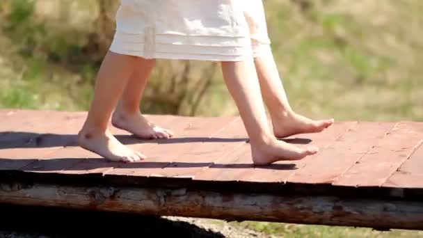Δύο κορίτσια δίδυμοι σε άσπρα λευκά φορέματα περπατούν σε μια ξύλινη γέφυρα στην ύπαιθρο μια καλοκαιρινή μέρα. Μέρη σώματος — Αρχείο Βίντεο