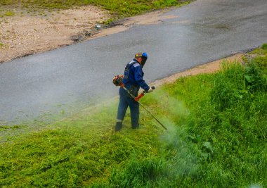 Bir adam, koruyucu bir üniformayla, benzin tırpanıyla çim biçiyor.