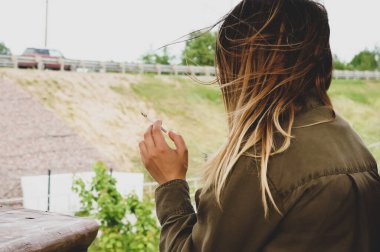 Kız elinde bir sigara tutuyor ve saçları rüzgarda dalgalanıyor.