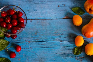 Mevsimlik kayısılar, cam bir kasede armut ve kiraz, mavi ahşap bir arka planda, mesajınız için yer var.