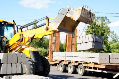 Traktör bir araba römorkuna kovayla beton bloklar yüklüyor.