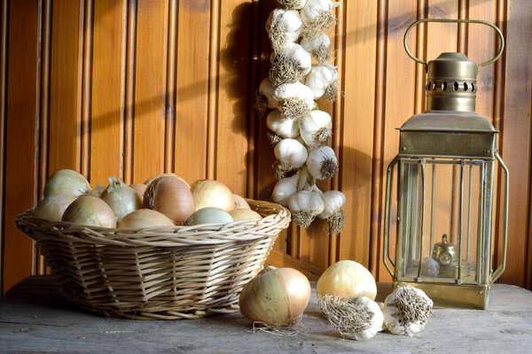 Onions on a basket, garlic braid hanging on wood wall and an oil lam