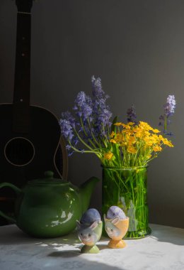 Vibrant spring still life with blue and yellow flowers in green glass vase, bunny egg cups with purple eggs, ceramic teapot, and guitar, Easter decor