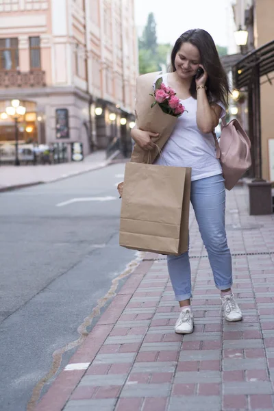 A woman with a bouquet and shopping speaks on the phone in the street.
